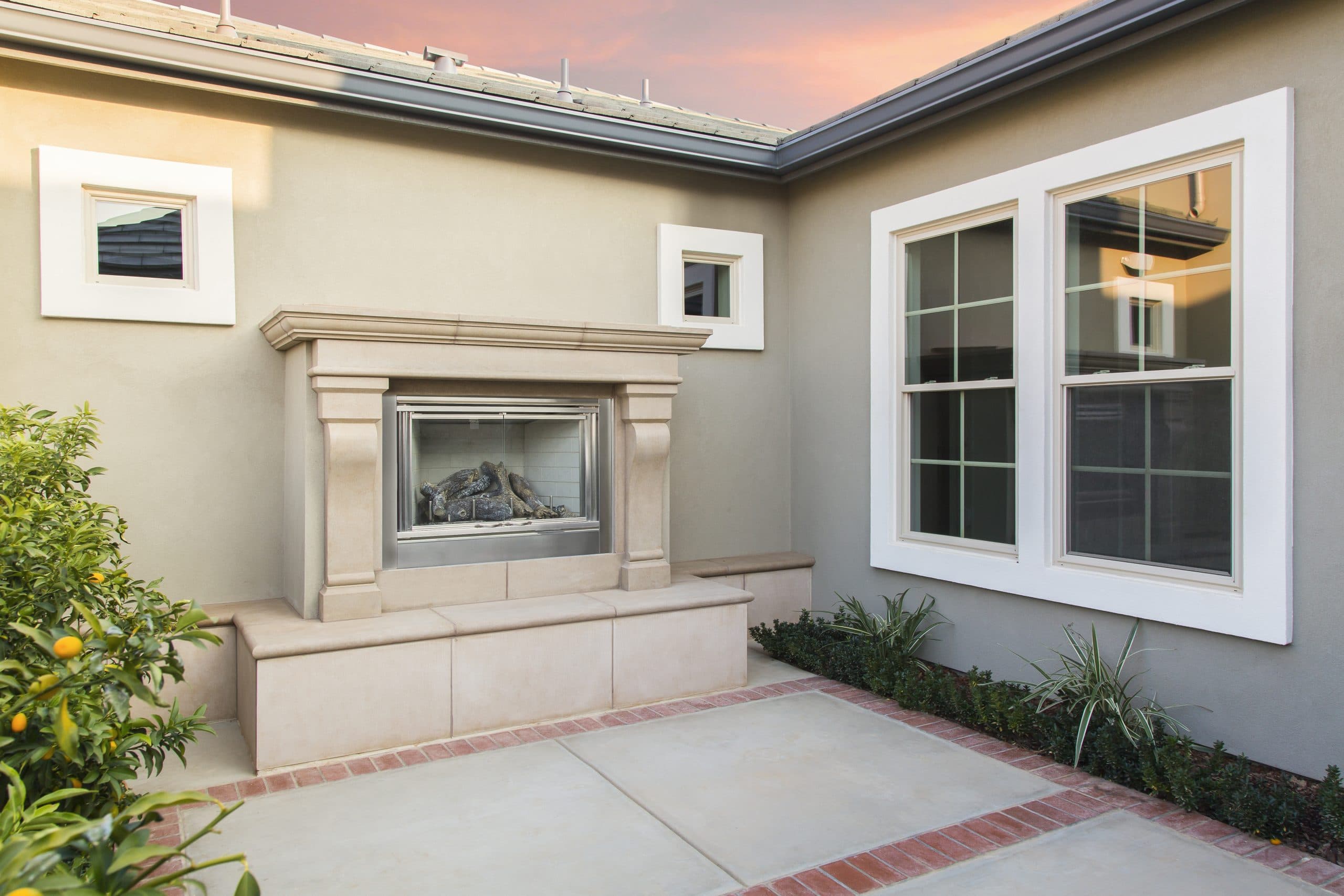 Stucco courtyard exterior at dusk — large white-frame Anlin grid-pane window beside an outdoor fireplace, with two square accent windows on the adjacent wall